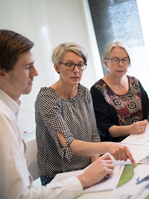 3 People in the middle of a meeting, they are looking at paperwork on the table and the woman in the middle is pointing at something and talking.